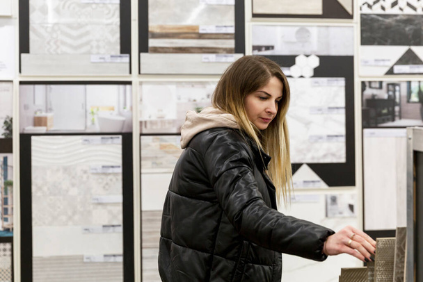 A young woman chooses ceramic tiles in a hardware store. Beautiful blonde in a black jacket. Construction, design and repair. - Fotografie, Obrázek
