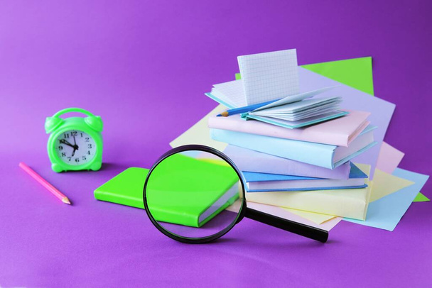 Books, magnifying glass, alarm clock and notepads, pastel colored paper, on the table, purple background, the concept of learning, education, back to school, holidays, reading - Photo, Image