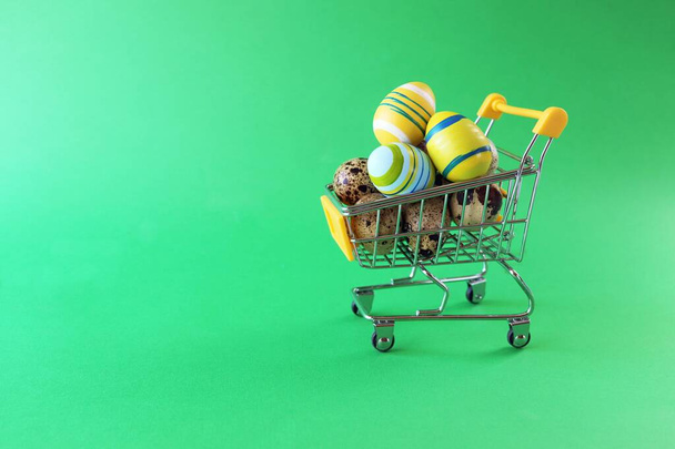 Frohe Ostern, Supermarkt-Spielzeugwagen gefüllt mit Wachteln und bunten Eiern, leuchtend grüner Hintergrund mit Kopierraum, Osterverkaufskonzept, traditionelle Dekoration für den Feiertag, Glückwunsch, Postkarte - Foto, Bild