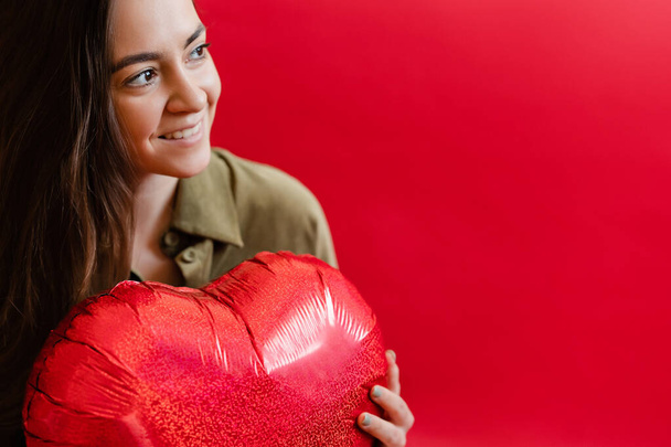 Šťastná dívka drží červený balón ve tvaru srdce. Fotografie usmívající se mladé dívky v lásce na červeném pozadí. Valentýn s kopírovacím prostorem. Dámský den - Fotografie, Obrázek