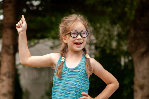 Young happy curious elementary school age girl, clever intelligent genius child student in big funny glasses has a brilliant idea, finger pointing up, eureka pose concept one person, outdoors portrait - Photo, Image