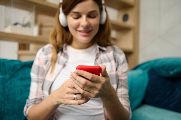 Focus on the hands of a blurred beautiful Caucasian woman using mobile phone, listening to the music in wireless headphones while relaxing on the couch at home, enjoying her quit pregnancy - Foto, imagen