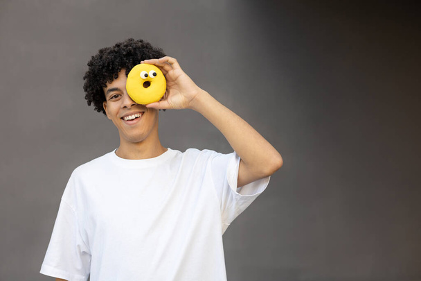 Fast food, dessert, unhealthy eating and people concept - Happy American guy holding a yellow donut in his hand instead of his eye, wearing a white T-shirt on a gray background with copy space - Zdjęcie, obraz