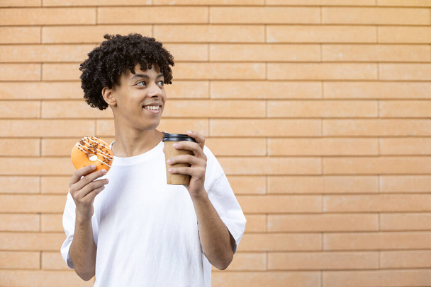 Dessert, leisure and people concept - American guy holding a cup of coffee and an orange glazed donut, looking away and wearing a white T-shirt on a brick wall background with copy space - Foto, Imagem