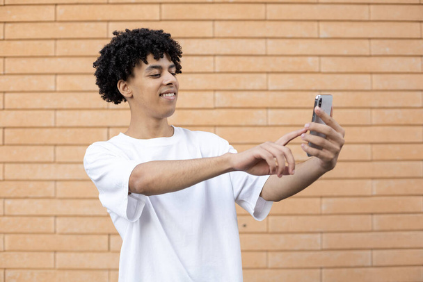 Technologie, Bildung und Menschen-Konzept - Ein glücklicher amerikanischer Mann hält ein Telefon in der Hand und dreht das Mikrofon mit dem Finger auf dem Bildschirm während eines Online-Treffens an einer Ziegelwand - Foto, Bild