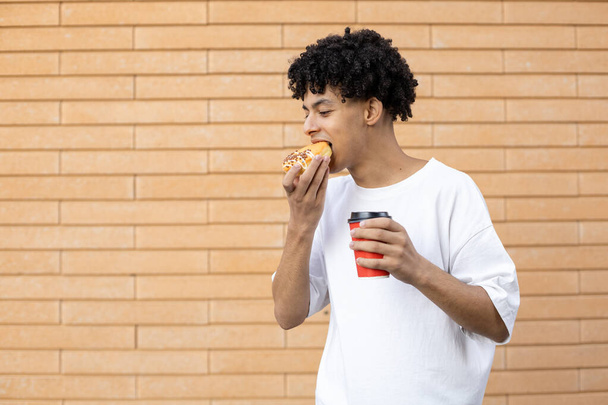 Dessert, bevande, stile di vita e concetto di persone - Un uomo afroamericano sorridente che tiene una tazza di caffè e morde una ciambella arancione, guarda e indossa una t-shirt bianca su uno sfondo muro di mattoni - Foto, immagini