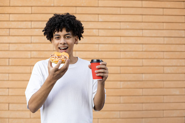 Sobremesa, bebidas, estilo de vida e conceito de pessoas - Um homem afro-americano feliz mordendo um donut laranja e segurando uma xícara de café, olhando para a câmera e vestindo uma camiseta branca em uma parede de tijolo  - Foto, Imagem