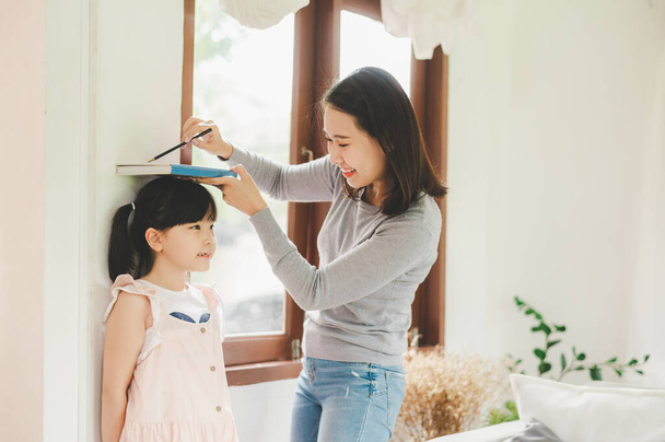 Happy Asian mother using book and pencil to measure height of little girl near wall at home - Photo, Image