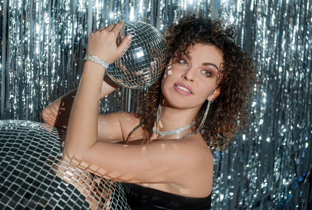 Beautiful young woman with curly hair in black body holding disco ball in hands. Woman posing on disco background with balls and sparkling texture - Photo, image