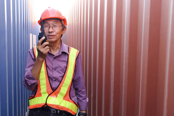Porträt eines älteren asiatischen Ingenieurs, der Sicherheitsweste und Helm trägt, Funkgeräte in der Hand hält und zwischen roten und blauen Containern auf dem Logistikcontainerplatz steht. - Foto, Bild