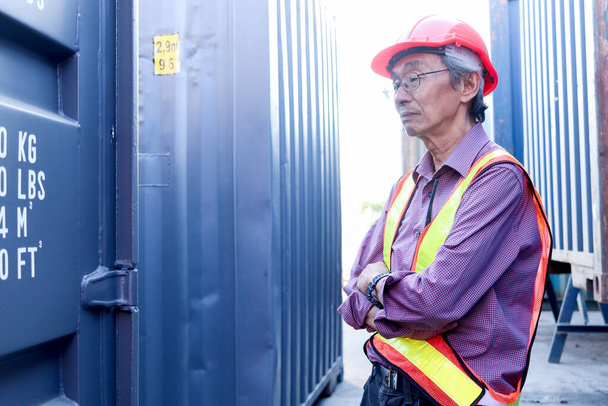 Портрет серьезного пожилого азиатского рабочего-инженера в спасательном жилете и шлеме, стоящего со скрещенными руками с голубыми контейнерами в качестве фона на логистической судоходной грузовой верфи. - Фото, изображение