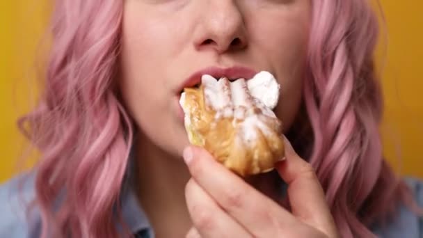 Close-up van aantrekkelijke vrolijke hongerige hongersnood meisje met roze haar kapsel genieten van bijten smakelijke zoete room cupcake geïsoleerd over gele achtergrond - Video