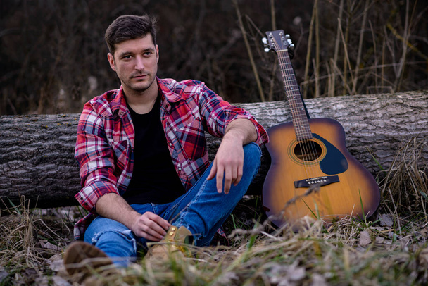 Yakışıklı genç bir adam, bir doğa parkındaki akustik gitarın yanındaki devrilmiş bir ağaca yaslanmış yerde oturuyor.. - Fotoğraf, Görsel