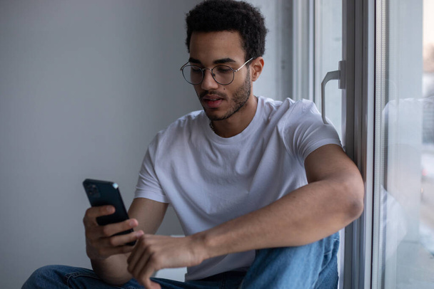 African American man in glasses and a white t-shirt uses a smartphone while sitting by the window. - Zdjęcie, obraz