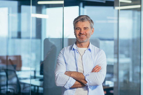 Portrait of a successful and happy gray-haired boss, a businessman in a modern office marveling at the camera and smiling with crossed arms - Zdjęcie, obraz