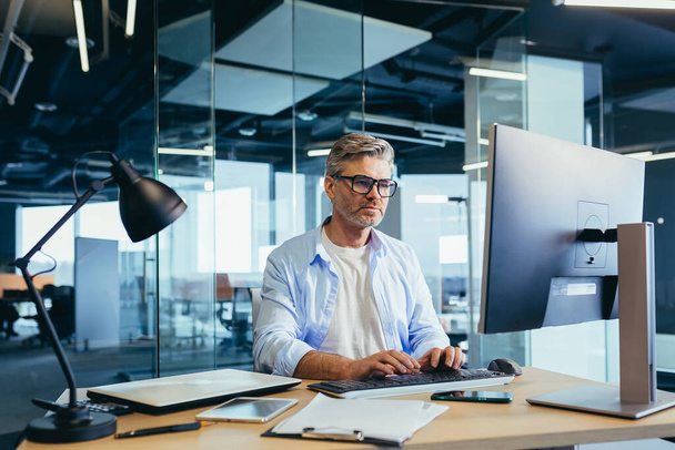 Successful senior boss with glasses, working on computer, businessman in shirt, in modern office at work - Photo, image