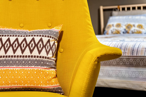 Schlafzimmerkonzept in den Farben gelb, beige und grau. Ein Teil des Sessels und des Kissens darauf in selektiver Fokussierung, das Bett im verschwommenen Hintergrund.  - Foto, Bild