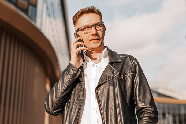Handsome man with glasses with a smartphone on the street of a big city. Businessman talking on the phone on urban background - Zdjęcie, obraz