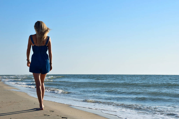 Una chica rubia en overoles de mezclilla está caminando por la playa de arena de mar. Vista trasera. A la derecha hay un lugar para insertar una inscripción - Foto, imagen