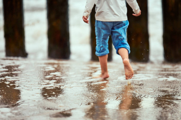Mały chłopiec w niebieskich spodniach biegający po wybrzeżu i bawiący się falami morskimi, nieostre rozmazane nogi. Mały chłopiec biegający po plaży przez plamy i krople wody morskiej, szczęśliwy pomysł letniego dzieciństwa - Zdjęcie, obraz