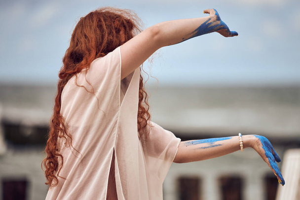Vörös hajú nő táncol a tengerparton, szabadtéri művészeti előadás. Vörös hajú lány, kék festékkel borított kezekkel, zenére táncol a tenger partján. Gyönyörű meditatív tánc a művészeti fesztiválon - Fotó, kép
