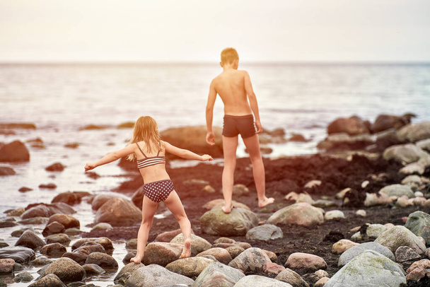 Young boy and girl on stone seacoast. Brother and his little sister rear back view, children playing catch on sea shore rocks coated with algae seaweed. Kids by sea, horizon background - Photo, Image