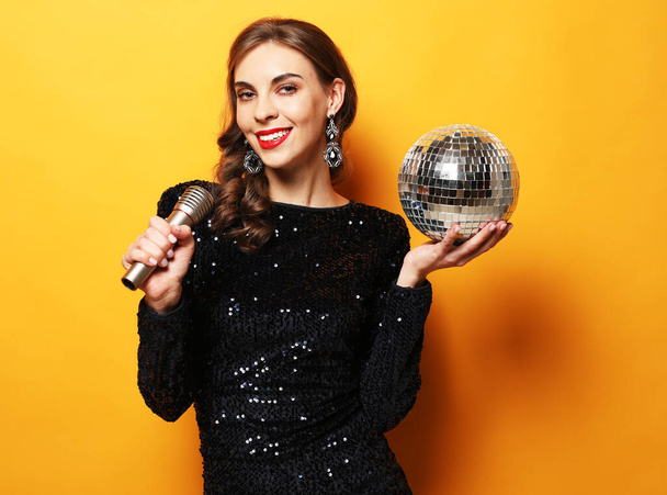 Young woman in evening dress holding microphone and disco ball. Brighrt make up and Wavy hairstyle. - 写真・画像