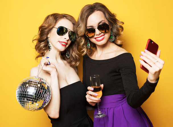 two fashionable young women holding disco ball, drinking champagne and taking a selfie. - Foto, afbeelding