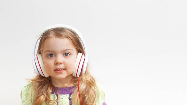 Весела маленька дівчинка 3 роки в навушниках на білому тлі з копіювальним простором. Музика або розваги онлайн
. - Фото, зображення
