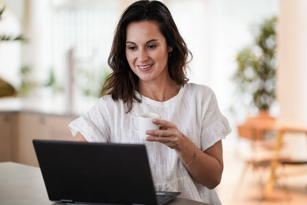 happy smiling remote working woman drinking coffee or espresso infront of a laptop or notebook on her work desk in her modern airy bright living room home office  - Photo, Image
