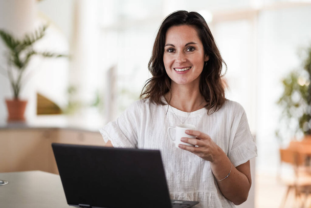 happy smiling remote working woman drinking coffee or espresso infront of a laptop or notebook on her work desk in her modern airy bright living room home office  - Fotoğraf, Görsel