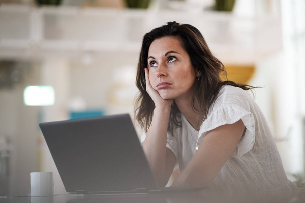 sad depressed remote working brown hair woman sitting disappointed with headache infront of a laptop or notebook on her work desk in modern airy bright living room home office  - Foto, immagini
