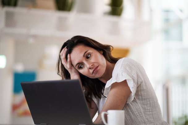 sad depressed remote working brown hair woman sitting disappointed with headache infront of a laptop or notebook on her work desk in modern airy bright living room home office  - Foto, afbeelding