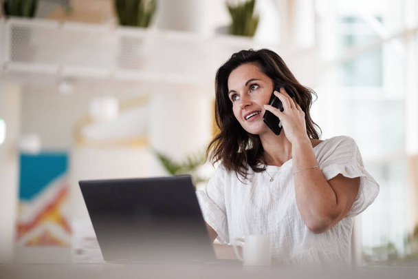 smiling remote working dark haired woman calling and chatting infront of a laptop or notebook in casual outfit on her work desk in her modern airy bright living room home office with her mobile phone - Foto, imagen