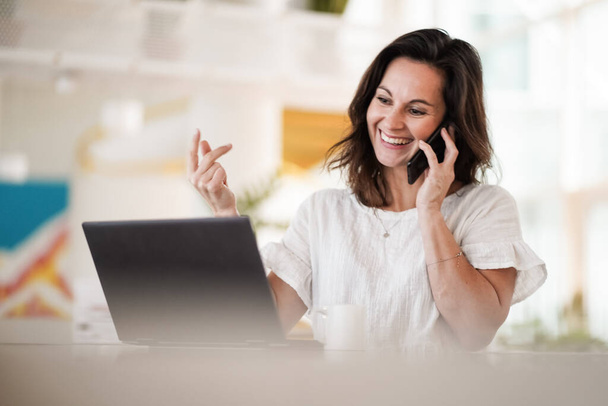 smiling remote working dark haired woman calling and chatting infront of a laptop or notebook in casual outfit on her work desk in her modern airy bright living room home office with her mobile phone - Photo, Image