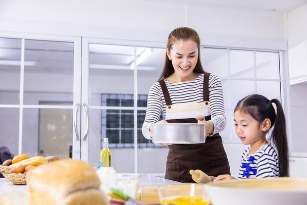 Ευτυχισμένη Ασιάτισσα μητέρα μαθαίνει στη μικρή της κόρη να ψήνει ψωμί στη μοντέρνα λευκή κουζίνα ενώ κοσκινίζει αλεύρι για ανάμιξη - Φωτογραφία, εικόνα