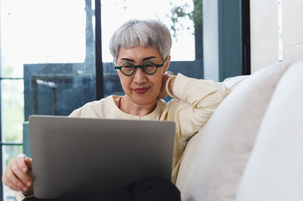 幸せなアジアのシニア女性グレーの髪はリビングルームのソファに座ってラップトップコンピュータを使用して眼鏡をかけます。ビデオチャットをする高齢女性をリラックスさせます。テクノロジーと古い時代の概念は. - 写真・画像