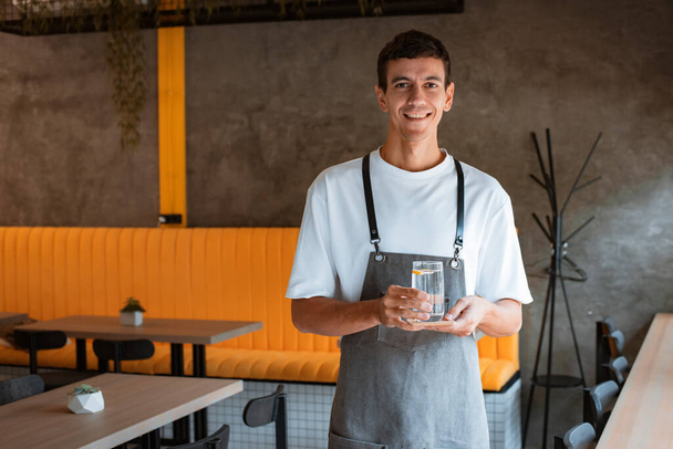 Young pleasant man cafe worker in apron serving table in local new stylish coffeehouse or coffee shop, waiter carefully placing glass of water and coaster on wooden table - Фото, изображение