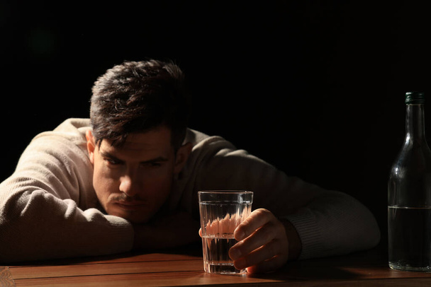 Addicted man with alcoholic drink at wooden table against black background, focus on glass - Photo, image