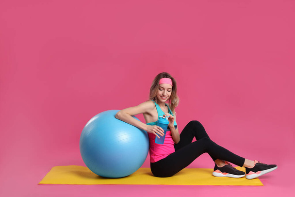 Beautiful woman sitting on yoga mat with fitness ball against pink background - 写真・画像
