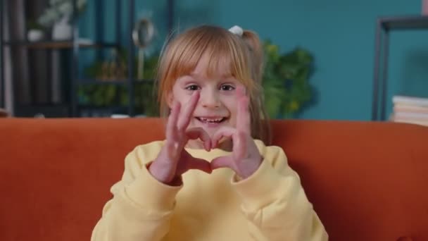 glimlachend kind meisje kind op home sofa kijken naar camera maakt hart gebaar demonstreert liefde teken - Video