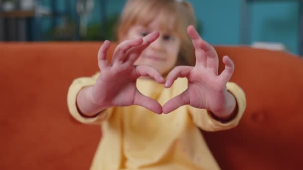 glimlachend kind meisje kind op home sofa kijken naar camera maakt hart gebaar demonstreert liefde teken - Video