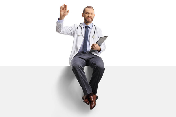 Young male doctor waving and sitting on a blank panel with a clipboard in his hands isolated on white background - Foto, Bild