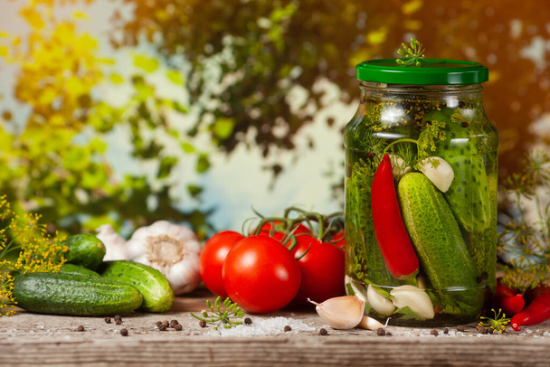 Pickled cucumbers in glass jars and spices and vegetables for preparation of pickles on background of green trees. - Photo, Image