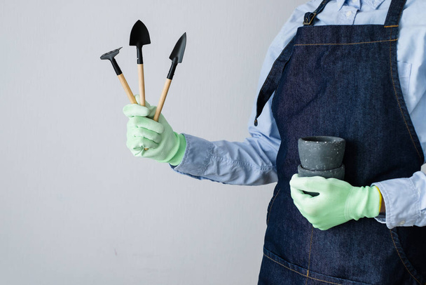 Mujer irreconocible con guantes sosteniendo herramientas en la mano y preparándose para trasplantar plantas de interior sobre un fondo blanco. Foto de alta calidad - Foto, Imagen