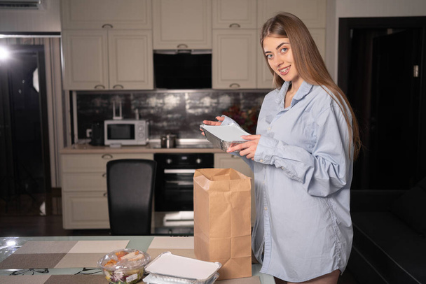 Μια όμορφη λευκή γυναίκα το βράδυ στο σπίτι βγάζει κουτιά μιας χρήσης για δείπνο με φαγητό στο τραπέζι από μια χάρτινη οικολογική τσάντα. Σκέψη για φαγητό. Αντιγραφή χώρου. - Φωτογραφία, εικόνα