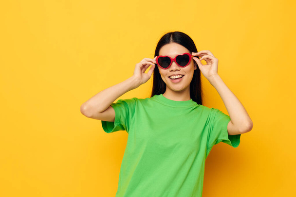 Charming young Asian woman in dark glasses in a green t-shirt posing modern yellow background unaltered - Fotó, kép