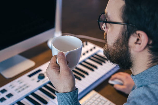 A man with a cup of coffee sits in front of a computer and a musical keyboard. - Fotoğraf, Görsel
