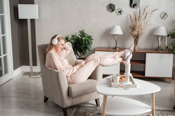 Beautiful middle-aged woman in home clothes and headphones sits in a chair with smartphones in hands and smiles. Pleasant online communication and relaxation at home. woman listens to music  - Fotoğraf, Görsel