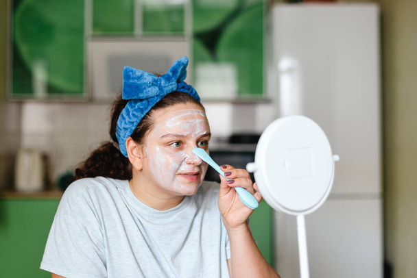 Ευτυχισμένη γυναίκα που εφαρμόζει ενυδατική μάσκα στο πρόσωπο για περιποίηση δέρματος με ειδικό ελαστικό πινέλο που φοράει μπλε φιόγκο στο κεφάλι κοιτάζοντας τον καθρέφτη. Αντιγηραντική. Αρχική διαδικασίες μακιγιάζ ομορφιά - Φωτογραφία, εικόνα
